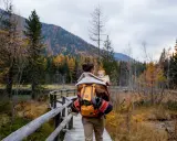 Ojciec z dzieckiem na plecach spaceruje drewnianą kładką pośród jesiennych lasów. Tatry z dzieckiem to piękna przygoda.