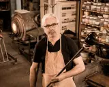 Un artisan verrier expérimenté, tablier en cuir, tient sa canne à souffler devant son four incandescent.
