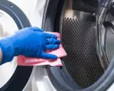 Mano con guante azul limpiando la goma de la lavadora con un paño rosa.