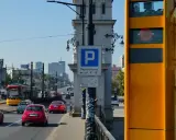 Żółty fotoradar stoi przy drodze, gdzie widać ruch samochodowy i tramwaj. Obok znak parkingu płatnego.