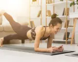 Uśmiechnięta kobieta w desce z nogą w górze, oglądająca trening na laptopie. To świetny sposób, jak zmotywować się do ćwiczeń w domu.