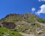 Bivacco Paolo e Nicola, un rifugio in legno ai piedi di montagne rocciose sotto un cielo azzurro.