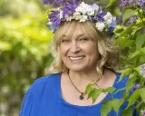 Sussie Eriksson med blomsterkrans i lila och vita blommor, en glad kvinna som påminner om hennes filmer och tv-program.