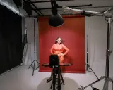 Modelka w pomarańczowej sukience pozuje na tle czerwonego tła. Wokół niej profesjonalne **światło studyjne** i sprzęt fotograficzny.