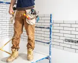 Pracownik na rusztowaniu, w spodniach roboczych i pasie z narzędziami, przygotowuje się do pracy. Widać białe płytki i gniazdka. Może to być etap, gdzie ujawnią się wady fugi epoksydowej.