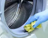 Mano con guante azul limpiando el borde de la lavadora con un paño amarillo. Un método natural para limpiar lavadora vinagre y bicarbonato.