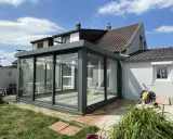Une véranda moderne en verre et métal, prête pour l'assurance. Le soleil brille sur le jardin et la maison.
