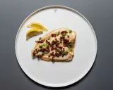Scholle Finkenwerder Art: gebratenes Fischfilet mit Speckwürfeln und Petersilie, garniert mit Zitronenspalten auf einem weißen Teller.