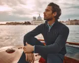 A man in mens dressy casual attire, a navy quarter-zip sweater over a blue collared shirt, sits on a boat with Venice in the background.