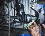 Mechanik wymienia uszkodzoną półoś, która powoduje wibracje. Widać gumową osłonę przegubu i zębaty wałek.