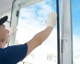 Un hombre con guantes blancos instala un vidrio simple en un marco de ventana blanco, con el cielo azul y nubes de fondo.