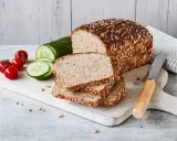 Frisch gebackenes Sonnenblumenbrot, zubereitet im Thermomix, liegt auf einem Holzbrett neben Tomaten und Gurkenscheiben.