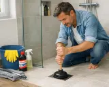 Un homme utilise une ventouse pour déboucher une douche. Des produits de nettoyage et des gants sont à proximité.