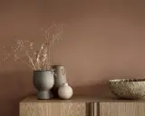 Tre vaser med torkade blommor och en flätad korg står på en träbyrå. En av vaserna är grå och har en rustik textur, en annan är beige med ett handtag och den minsta är rund och ljus.