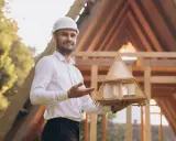 Architekt mit Helm hält Modellhaus, um Holzkonstruktion zu planen. Im Hintergrund Rohbau eines Hauses.