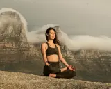 Kvinna i lotusställning på en klippa, med berg och moln i bakgrunden. Hon utövar buddhistisk meditation för inre lugn.