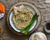 Hummus de berenjena cremoso, decorado con sésamo y espárragos, acompañado de pan de pita y crudités.
