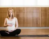 Gravid kvinna med slutna ögon sitter i lotusställning på en yogamatta. Hon håller sin mage, en lugn stund av gravid yoga.