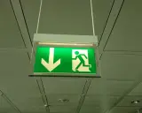 Zielony znak oświetlenia awaryjnego i ewakuacyjnego z ikoną strzałki w dół i biegnącej postaci.