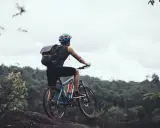 Rowerzysta z plecakiem na górskim szlaku, zastanawia się: gravel czy szosa? W tle zielone wzgórza i pochmurne niebo.