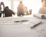 Na stole leżą plany budowlane, cyrkiel, linijka i długopis. W tle widać ludzi dyskutujących o zmianach w projekcie budowlanym bez zgody projektanta.