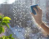 Hand mit Handschuh putzt Fenster mit Schwamm. Tropfen und Blasen auf der Scheibe deuten auf das Ziel hin: Fenster putzen ohne Schlieren.