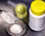 Ein Löffel mit weißem Pulver, bereit für die Reaktion mit Essig. Daneben ein Behälter mit Natron und eine Flasche mit Flüssigkeit.