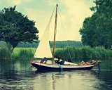 Un petit type de bateau à voile navigue sur un canal bordé d'arbres. Une personne se repose sur le pont, une autre est assise.
