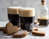 Två glas med mörk kaffedrink med skummande topp, omgivna av hjärtformade pepparkakor.