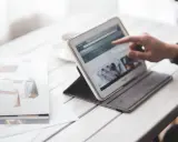 Dłoń wskazuje na ekran tabletu, który wyświetla stronę internetową. Obok leży otwarta książka z grafikami. W tle widać filiżankę. To idealne miejsce do pracy nad projektem, gdzie każdy element, nawet **padding CSS**, ma znaczenie.