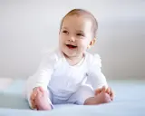 Uśmiechnięte dziecko w białym ubranku siedzi z podparciem. Od kiedy można sadzać dziecko z podparciem? Od momentu, gdy stabilnie trzyma główkę.