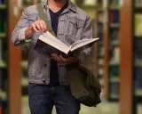 Student w dżinsowej kurtce przegląda książkę w bibliotece. W tle widać regały z książkami.