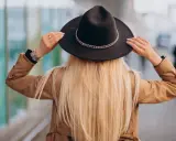 Blondynka w beżowym płaszczu i czarnym kapeluszu z ozdobnym łańcuszkiem. Pokazuje, jak nosić kapelusz, poprawiając go na głowie.