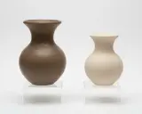 Två oglasade vaser i blålera keramik, en mörkbrun och en ljusbeige, står på genomskinliga piedestaler.
