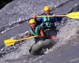 Dwóch uśmiechniętych ludzi w kaskach i kamizelkach pokonuje rwący nurt rzeki na pontonie. To właśnie rafting – emocjonująca przygoda!