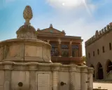 Fontana storica nel centro di Rimini, con edifici antichi sullo sfondo e cielo azzurro.