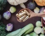 Łyżka z kapsułkami – witaminy na energię. W tle świeże warzywa: brokuły, brukselka, kapusta pak choi.