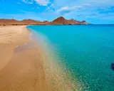La **playa de los Genoveses** se extiende con arena dorada y aguas cristalinas de color turquesa, bajo un cielo azul con nubes blancas y montañas áridas al fondo.
