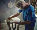 Artisan masqué ponçant une planche de surf blanche, étape cruciale avant de faire de l'époxy.
