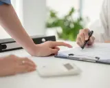 Podpisanie umowy zlecenia. Dwie osoby analizują dokumenty, jedna wskazuje palcem na tekst, druga trzyma długopis.