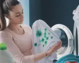 Mujer decepcionada revisa ropa blanca desteñida de azul, con manchas verdes, junto a una lavadora.