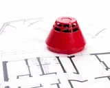 Roter Rauchmelder auf Bauplänen. Ein Beispiel für baulichen Brandschutz, der Sicherheit in Gebäuden gewährleistet.