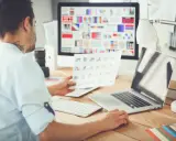 UX UI designer pracuje przy biurku, analizując wydruki i ekran komputera z paletą barw.