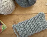 Ovillos de lana gris y beige sobre madera. Un trozo de tejido con puntos de lana muestra la textura.
