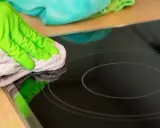 Grüne Handschuhe wischen mit einem Tuch über das Induktionskochfeld, um es zu reinigen.