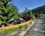 Un ruscello scorre a Santo Stefano di Cadore, fiancheggiato da case, alberi e un'area con gazebo e tavoli all'aperto.