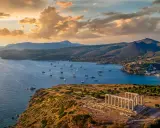 Rovine del Tempio di Poseidone a Capo Sounion, con barche a vela che navigano nel mare blu sotto un cielo dorato al tramonto.