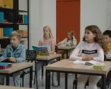 Dzieci siedzą w klasie, skupione na lekcji. Chłopiec w dżinsowej kurtce, dziewczynka z książką po angielsku, a z tyłu inne dzieci. Wszyscy mieli nadzieję, że lekcja szybko się skończy.