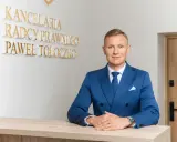 Mężczyzna w niebieskim garniturze przy biurku, za nim napis "KANCELaria Radcy Prawnego Paweł Tołoczko".