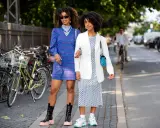 Two women showcase their spring chic dress code. One wears a blue plaid dress and chunky boots, the other a white blazer over a patterned dress and sneakers.